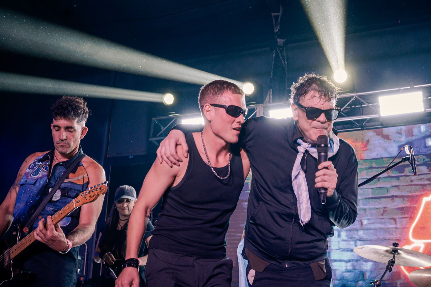 El cantante al micrófono, abrazado a un músico, al frente del escenario
